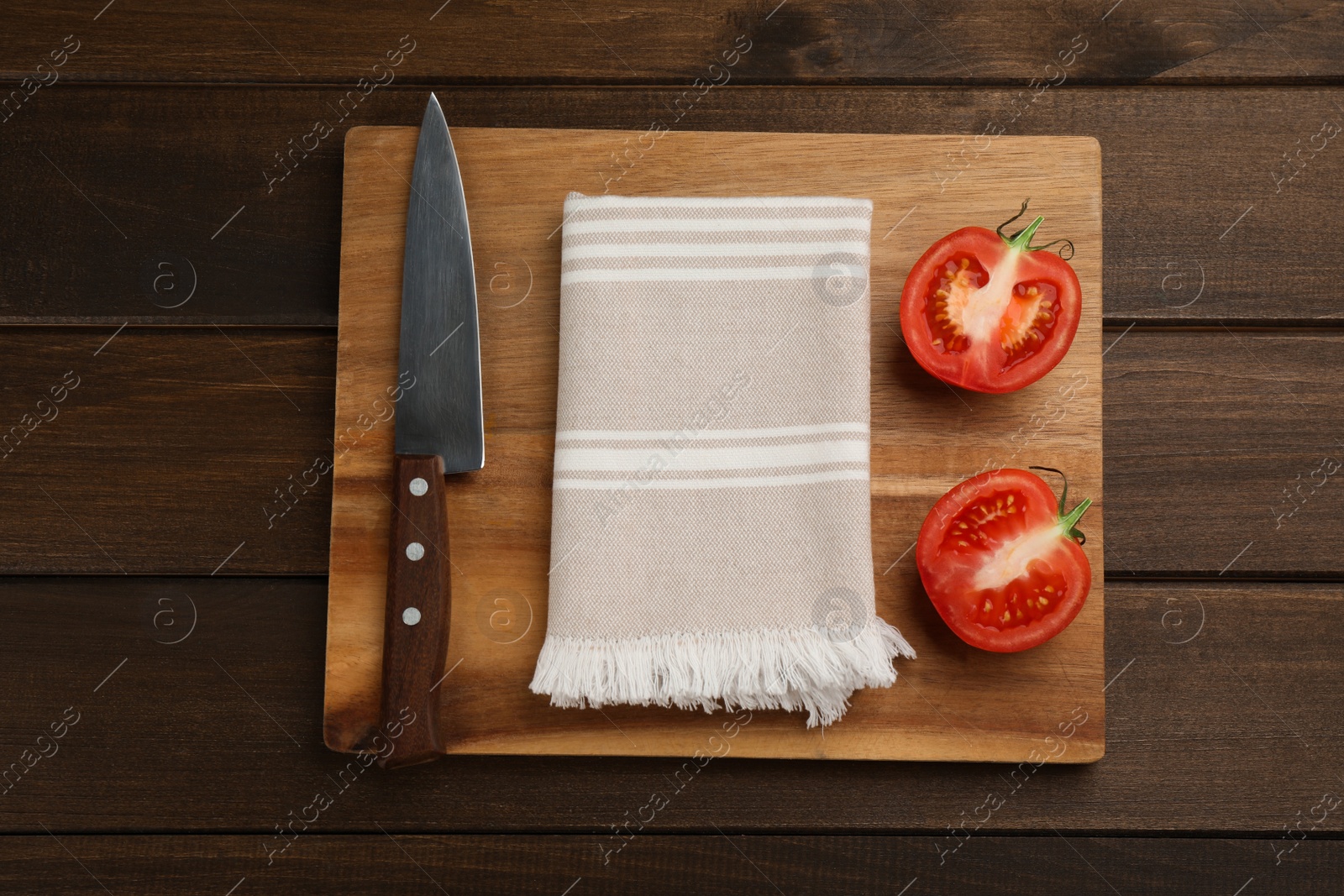 Photo of Cutting board with kitchen towel, halved tomato and knife on wooden table, top view