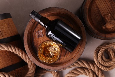 Photo of Whiskey with ice cubes in glass, bottle, wooden barrels and rope on grey marble table, flat lay