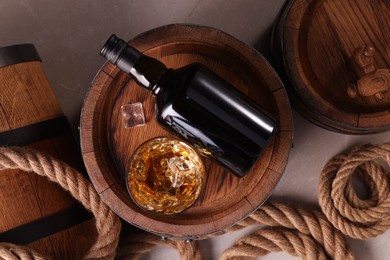 Whiskey with ice cubes in glass, bottle, wooden barrels and rope on grey marble table, flat lay