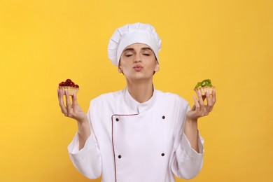 Professional confectioner in uniform with delicious tartlets on yellow background