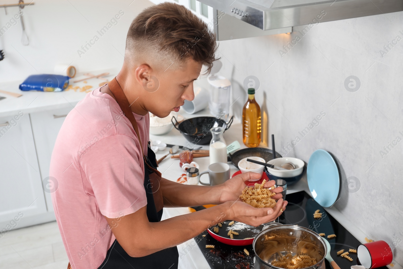 Photo of Man putting pasta into saucepan in messy kitchen. Many dirty dishware and utensils on stove and countertop