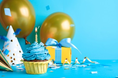 Photo of Delicious birthday cupcake with candle near gift box and party hats on light blue background, space for text