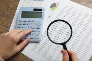 Photo of Woman looking at accounting document through magnifying glass while using calculator at wooden table, closeup