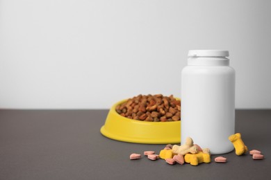 Bottle with vitamins and dry pet food in bowl on grey table, space for text