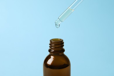 Dripping cosmetic oil from pipette into bottle on light blue background, closeup