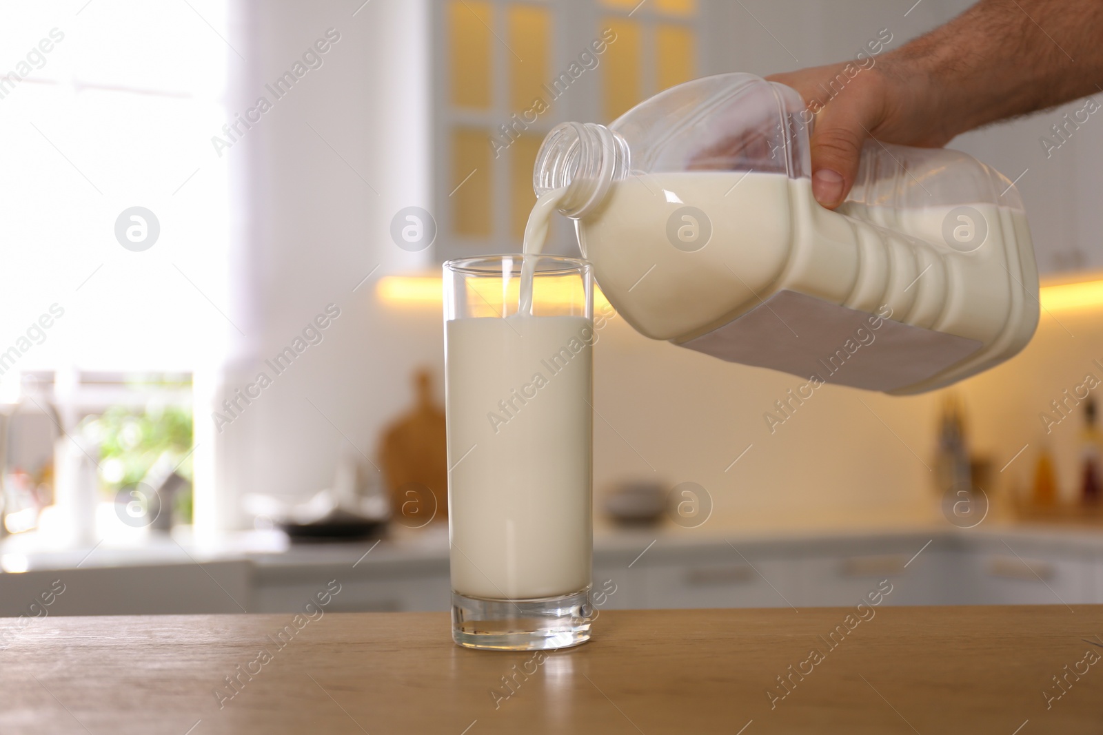 Photo of Man pouring milk from gallon bottle into glass at wooden table in kitchen, closeup