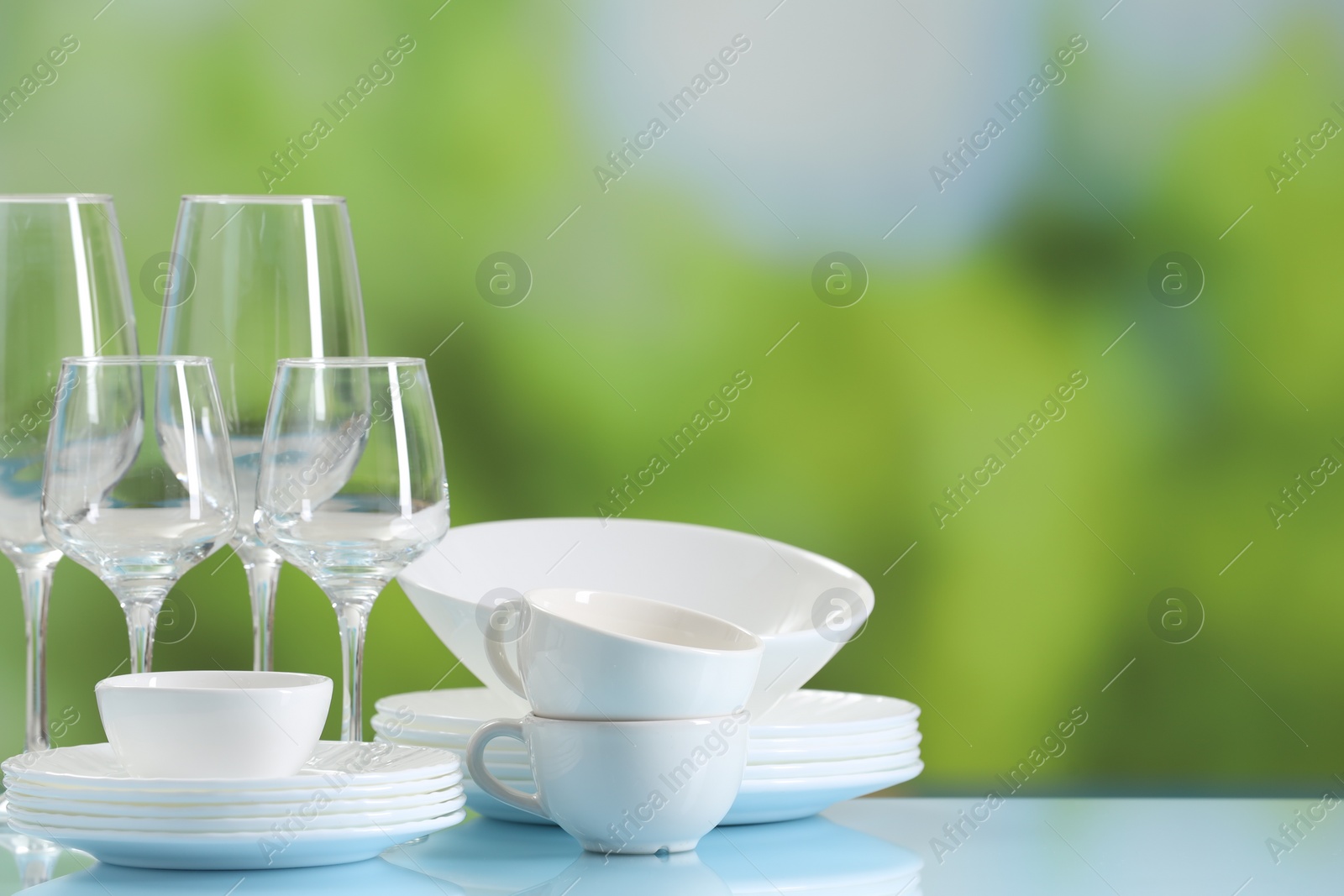 Photo of Set of many clean dishware and glasses on light blue table against blurred green background. Space for text