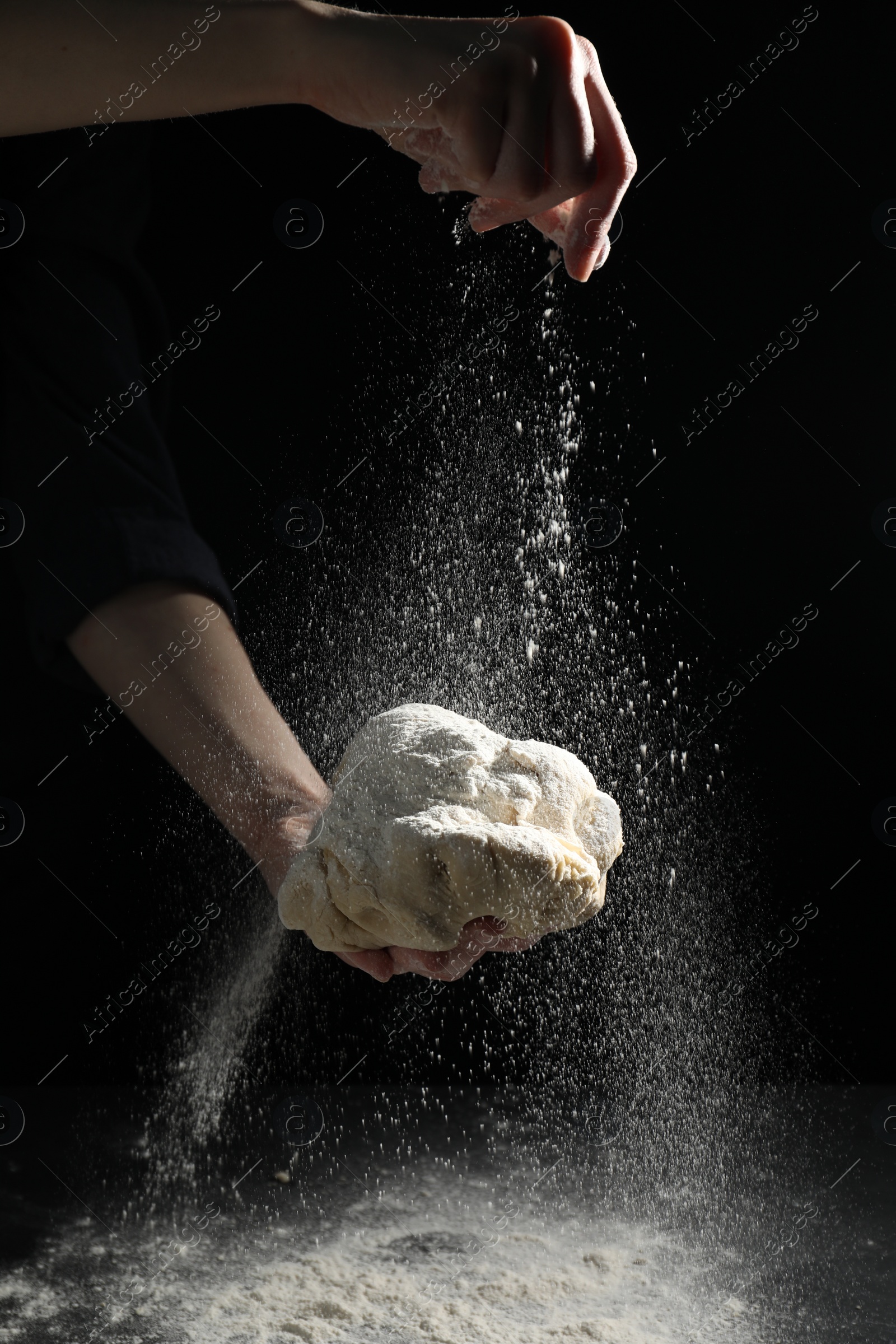 Photo of Making bread. Woman sprinkling flour over dough at table on dark background, closeup