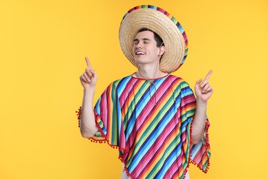 Photo of Young man in Mexican sombrero hat and poncho pointing at something on yellow background