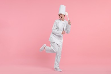 Happy professional confectioner in uniform holding whisk and spatula on pink background