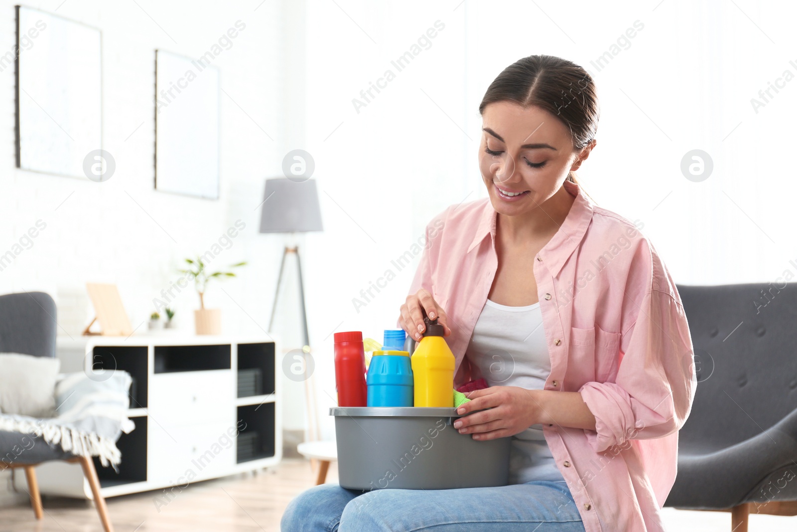 Photo of Portrait of young woman with basin of detergents in living room. Cleaning service