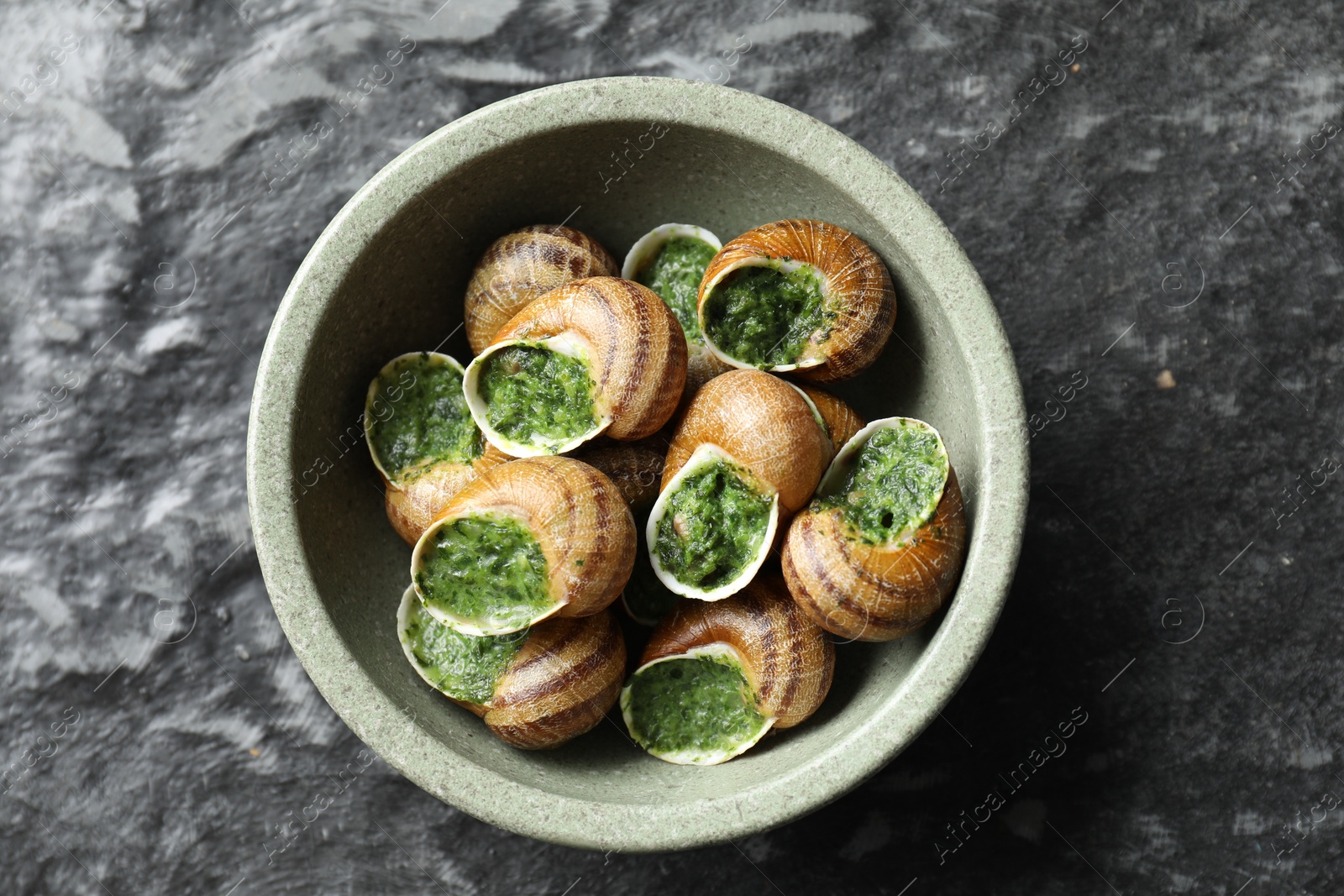 Photo of Delicious cooked snails in bowl on grey textured table, top view