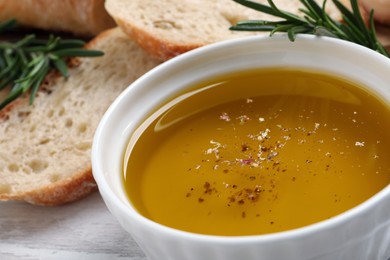 Photo of Bowl of fresh oil, bread and rosemary on white wooden table, closeup