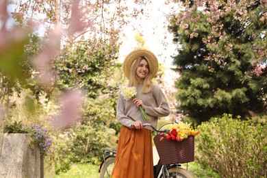 Beautiful young woman with bicycle and flowers in park on pleasant spring day