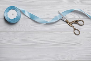 Pair of scissors with light blue satin ribbon on white wooden table, flat lay. Space for text