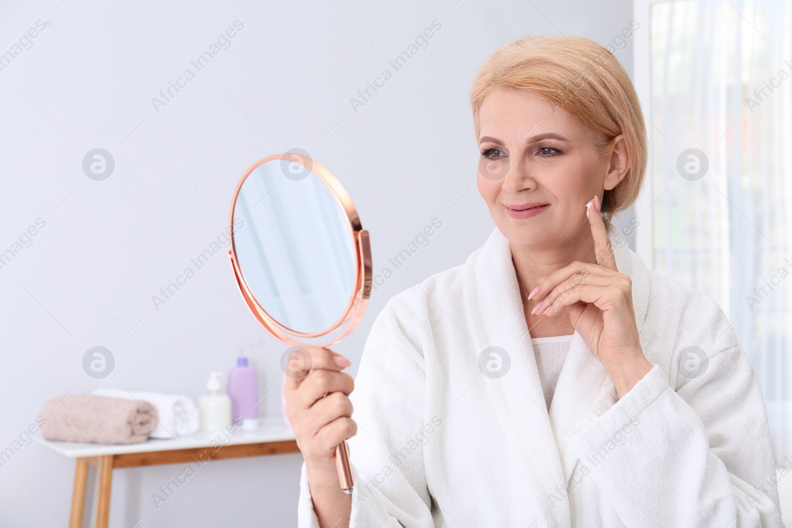 Photo of Mature woman with mirror applying face cream at home