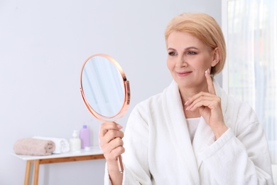 Mature woman with mirror applying face cream at home