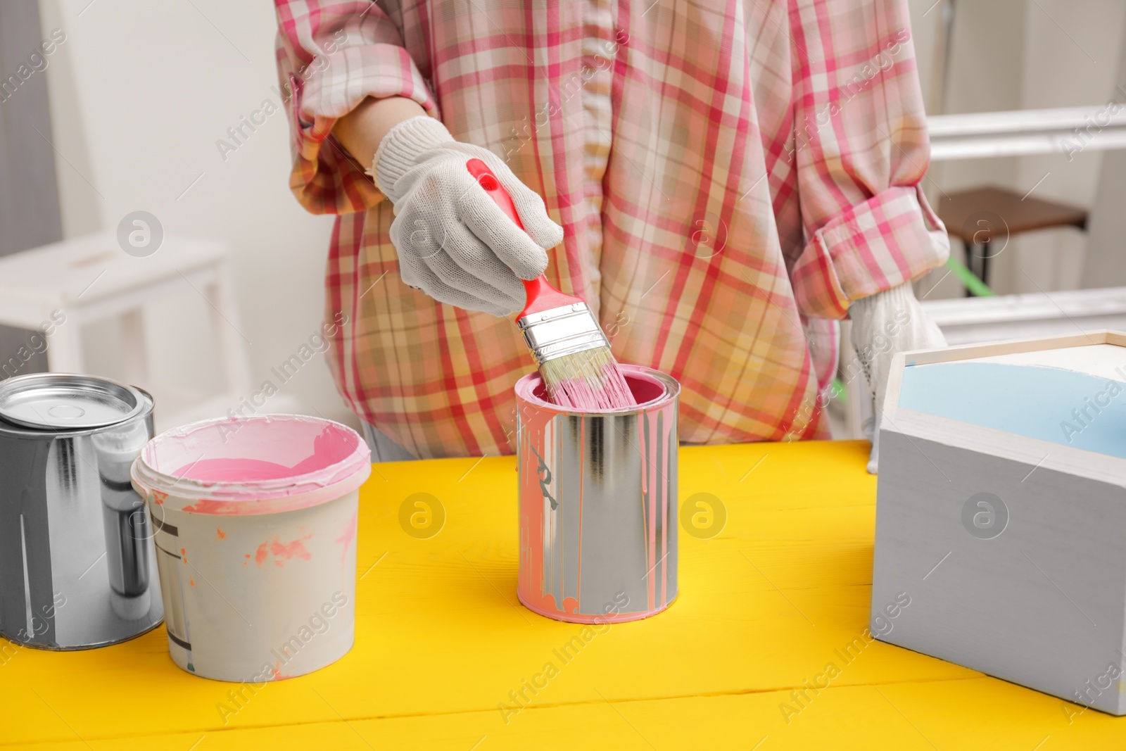 Photo of Woman dipping brush into can of pink paint at yellow wooden table indoors, closeup