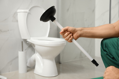 Photo of Professional plumber holding plunger near toilet bowl in bathroom, closeup