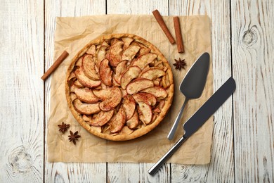 Photo of Delicious apple pie, cinnamon, spatula and knife on white wooden table, flat lay
