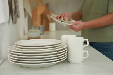 Woman wiping plate with towel at white table in kitchen, focus on dishware