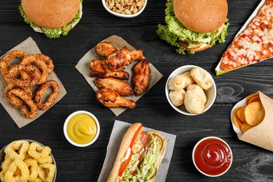 Chicken wings, onion rings and other fast food on black wooden table, flat lay