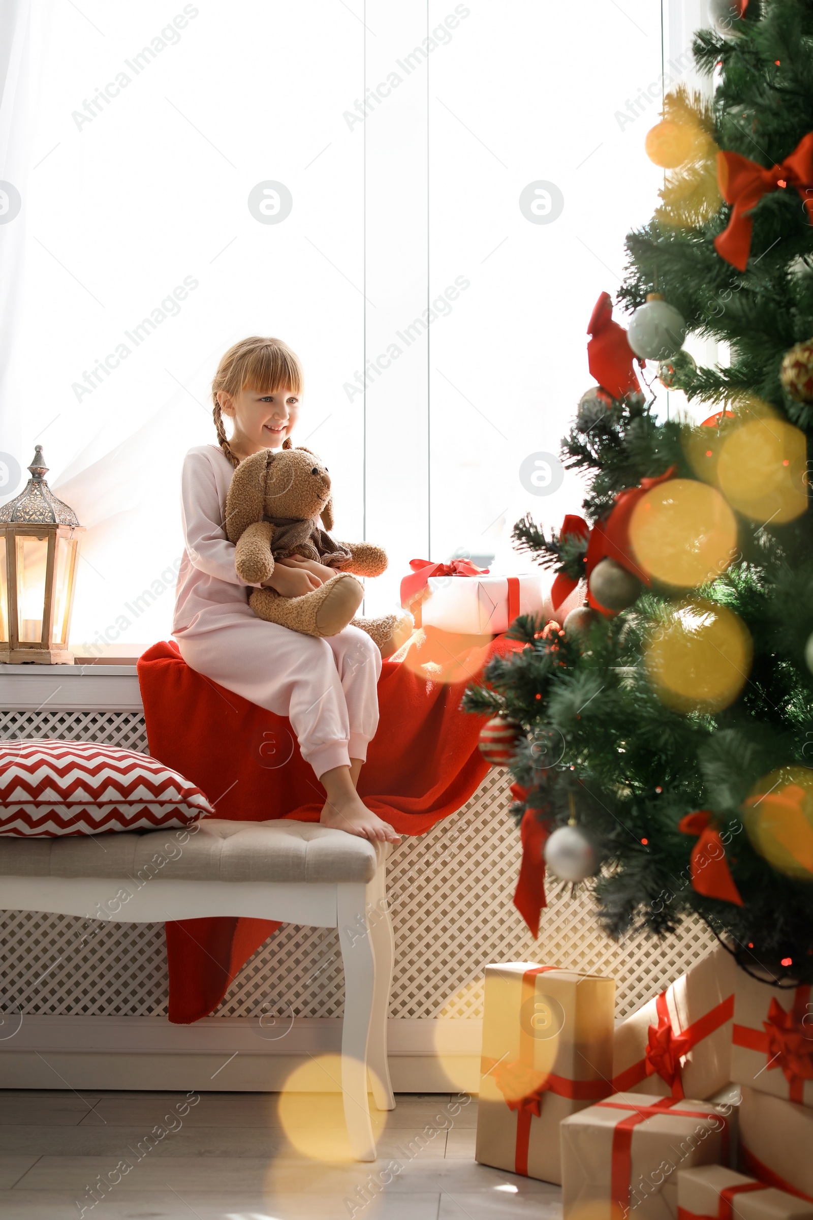 Photo of Cute little child with toy rabbit sitting on windowsill near Christmas tree at home