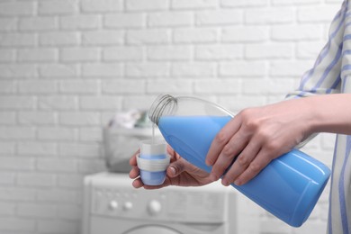 Woman pouring fabric softener from bottle into cap for washing clothes indoors, closeup