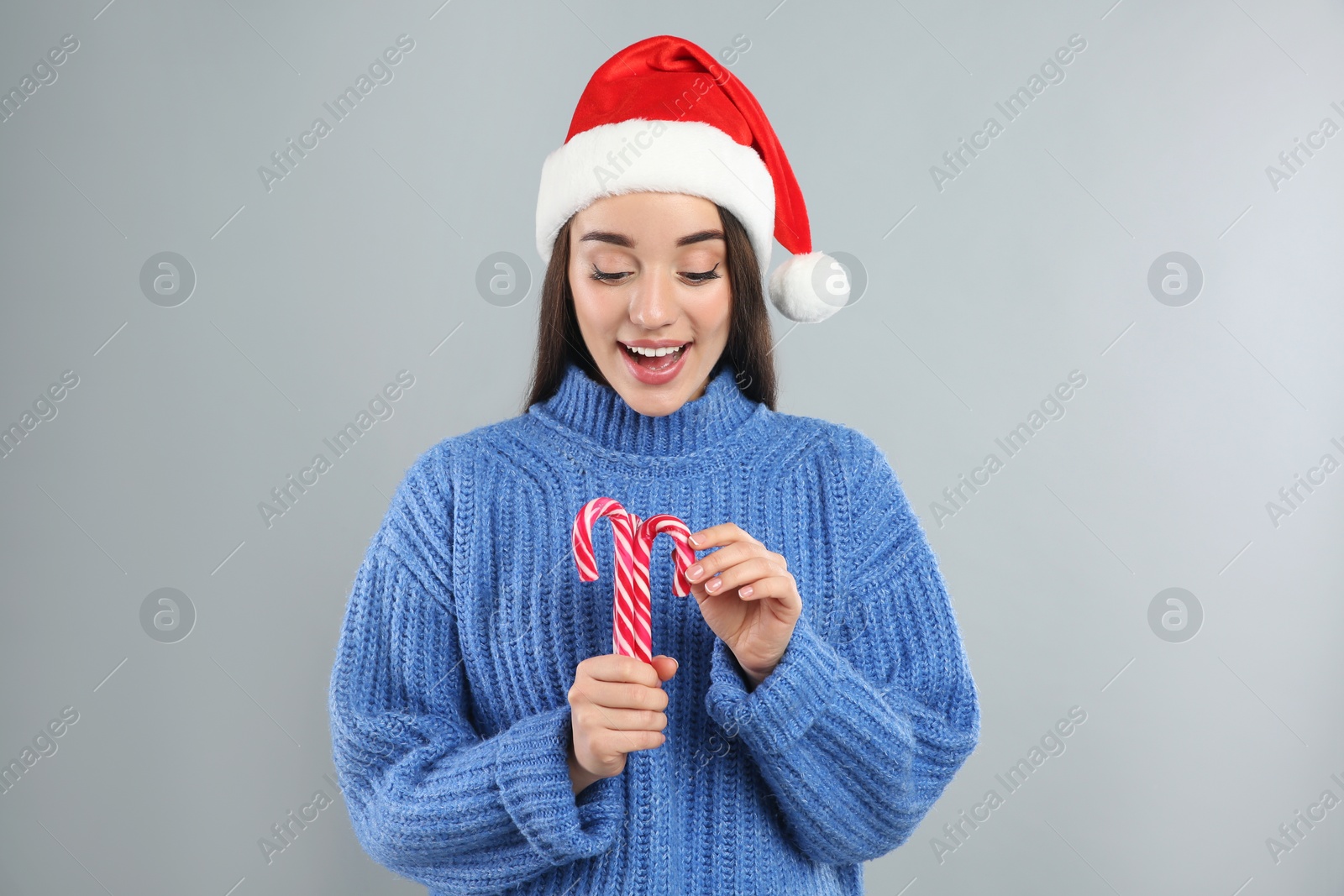 Photo of Young woman in blue sweater and Santa hat holding candy canes on grey background. Celebrating Christmas