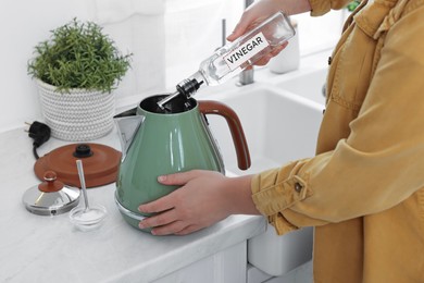Photo of Woman pouring vinegar from bottle into electric kettle in kitchen, closeup
