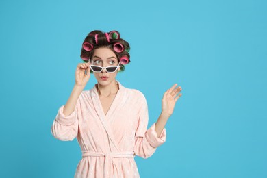 Photo of Emotional young woman in bathrobe with hair curlers and sunglasses on light blue background