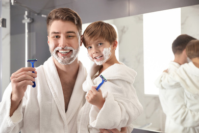 Photo of Dad and son with shaving foam on faces holding razors in bathroom