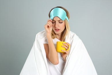 Young woman in sleeping mask wrapped with blanket holding cup on grey background