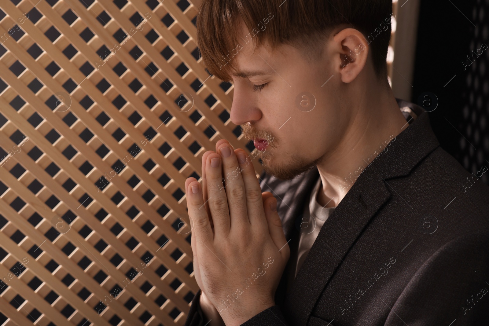 Photo of Man praying during confession near wooden window in booth. Space for text