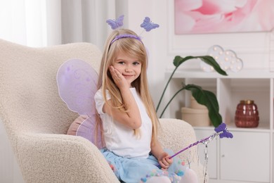 Cute little girl in fairy costume with violet wings and magic wand in armchair at home
