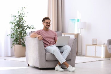 Photo of Handsome young man with laptop in armchair at home. Space for text