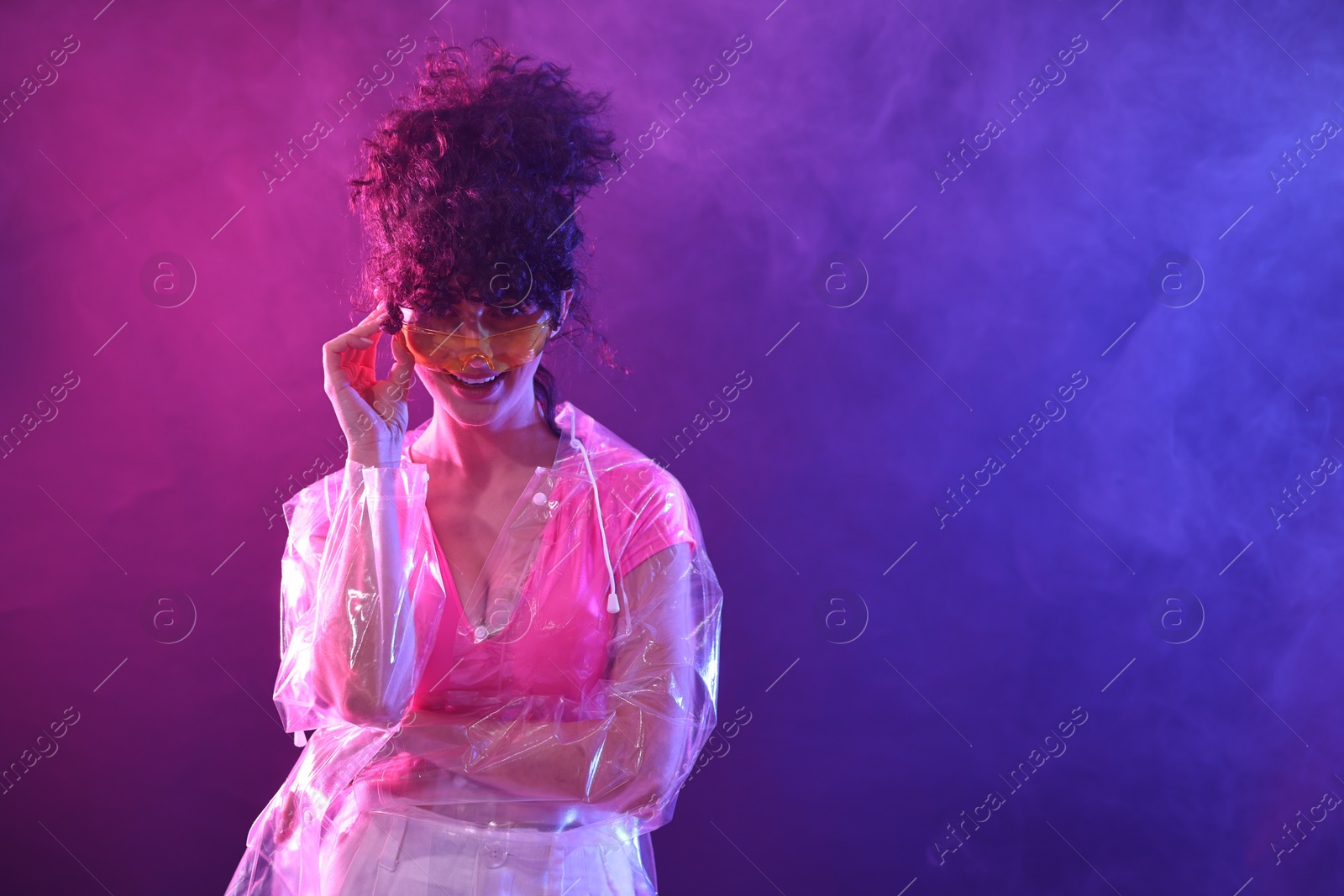 Photo of Beautiful young woman in transparent coat and sunglasses posing on color background in neon lights and smoke. Space for text