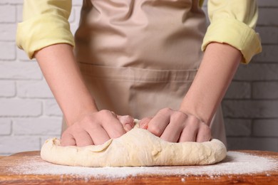 Photo of Woman kneading dough at wooden table near white brick wall, closeup
