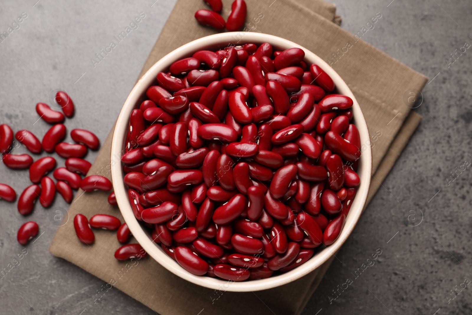 Photo of Raw red kidney beans in bowl and napkin on grey table, flat lay