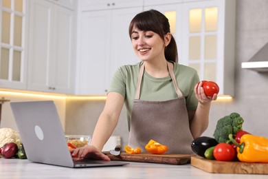 Photo of Happy young housewife using laptop while cooking at white marble table in kitchen