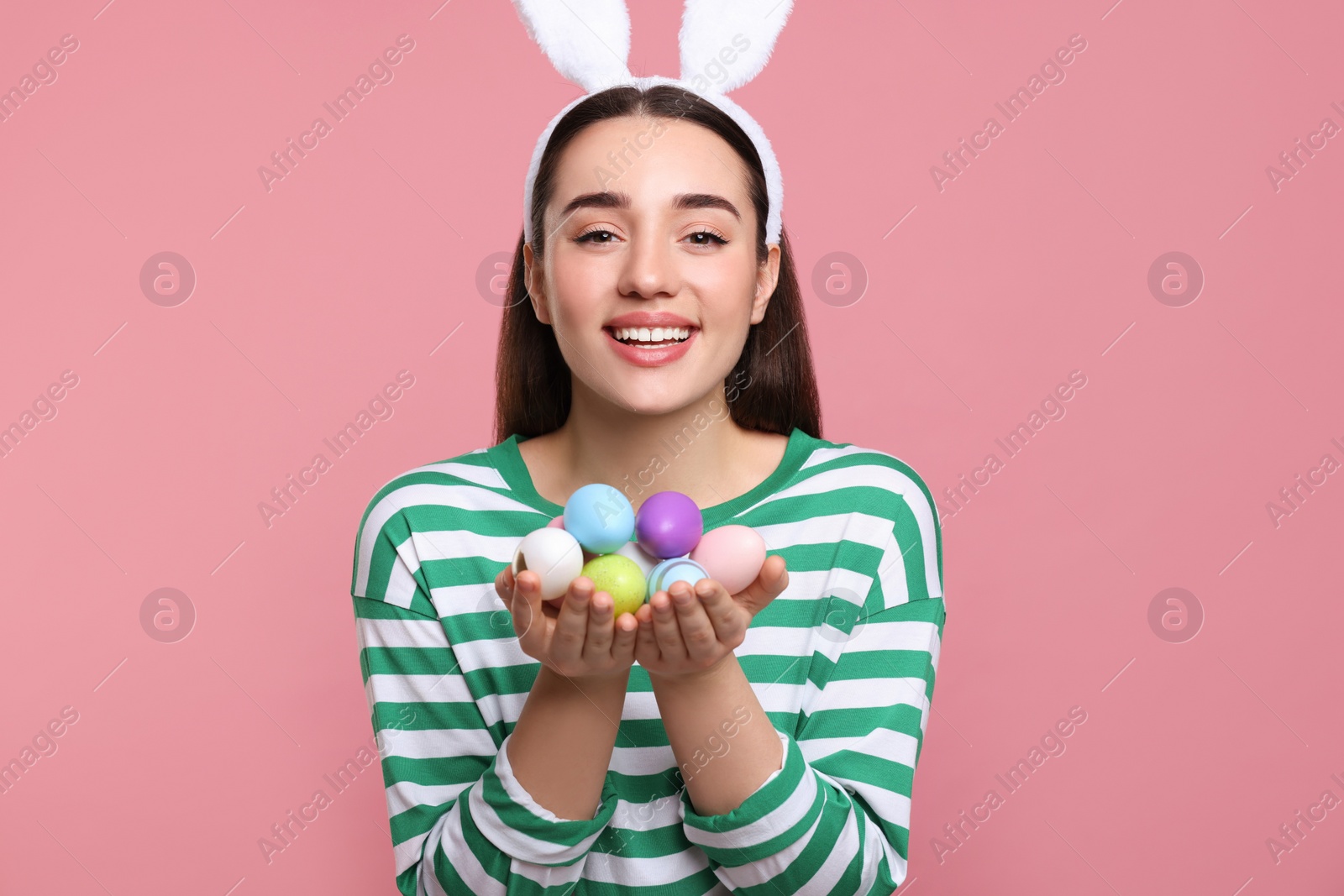Photo of Happy woman in bunny ears headband holding painted Easter eggs on pink background