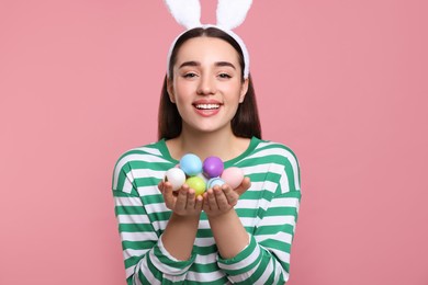 Happy woman in bunny ears headband holding painted Easter eggs on pink background