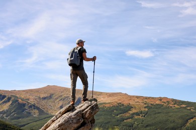 Photo of Man with backpack and trekking poles on rocky peak in mountains, back view