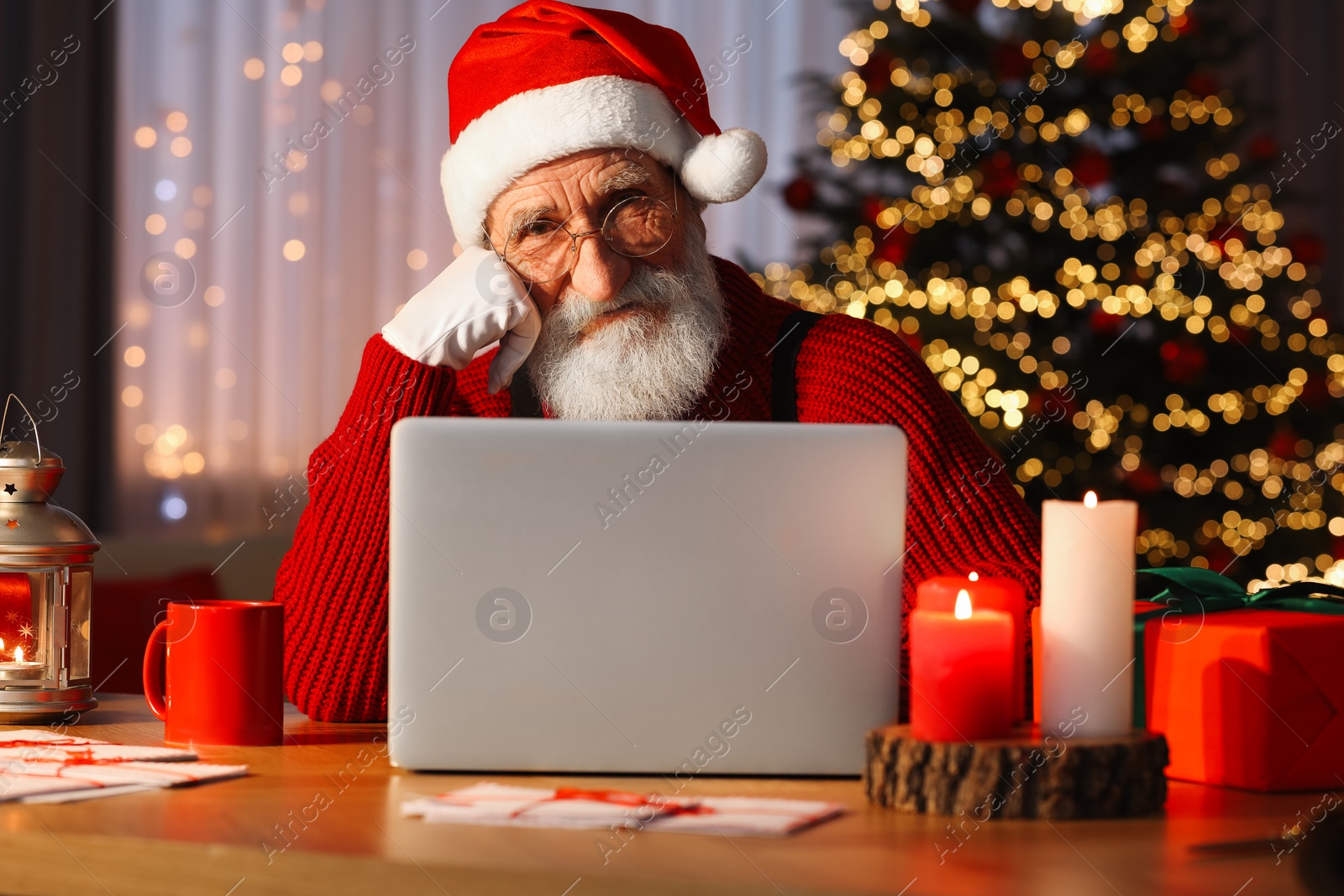 Photo of Santa Claus using laptop at his workplace in room with Christmas tree