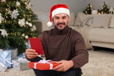 Photo of Happy man in Santa hat with Christmas gift and greeting card at home