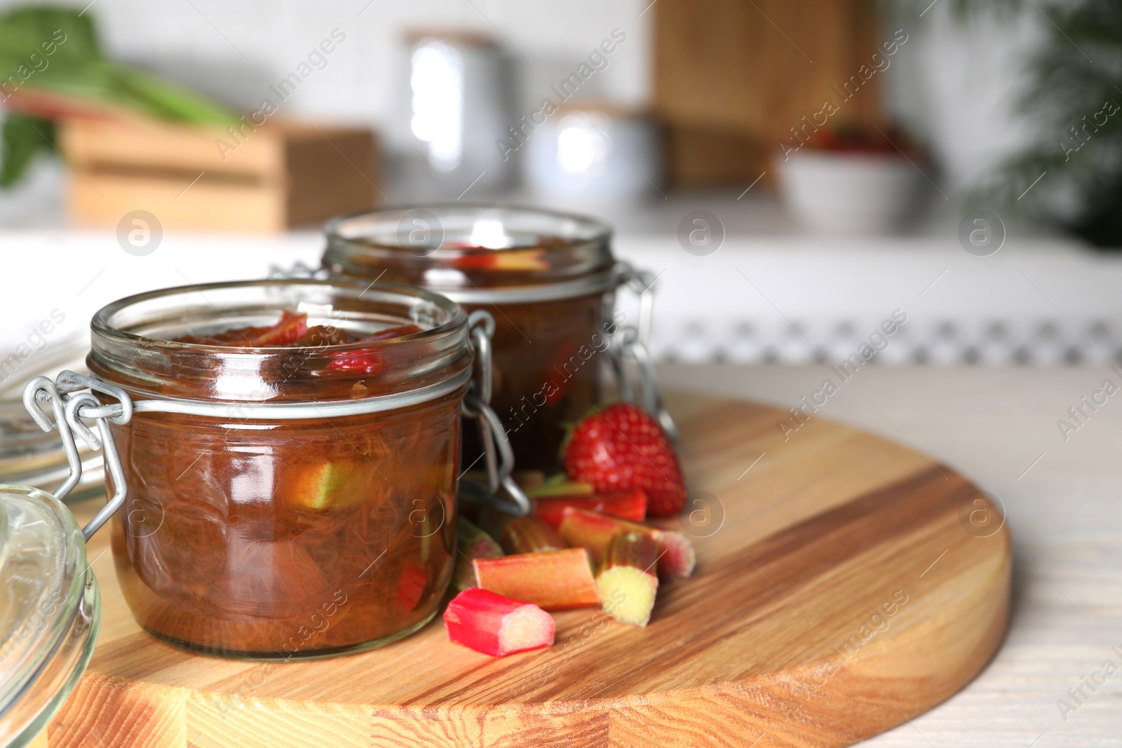 Photo of Jars of tasty rhubarb jam, cut stems and strawberry on white table. Space for text