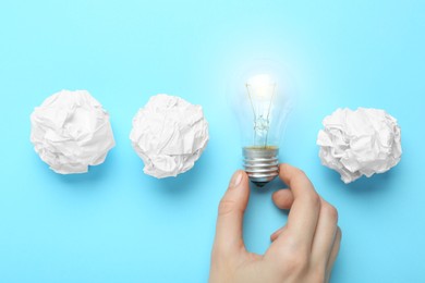 Photo of Woman holding lightbulb among paper balls on light blue background, top view. Idea concept