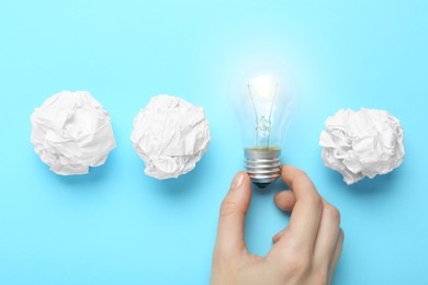 Woman holding lightbulb among paper balls on light blue background, top view. Idea concept