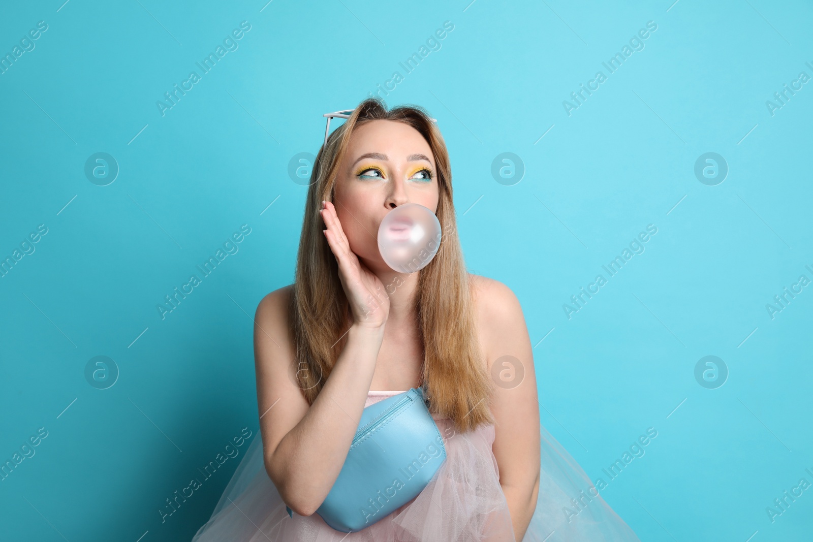 Photo of Fashionable young woman with bright makeup blowing bubblegum on light blue background