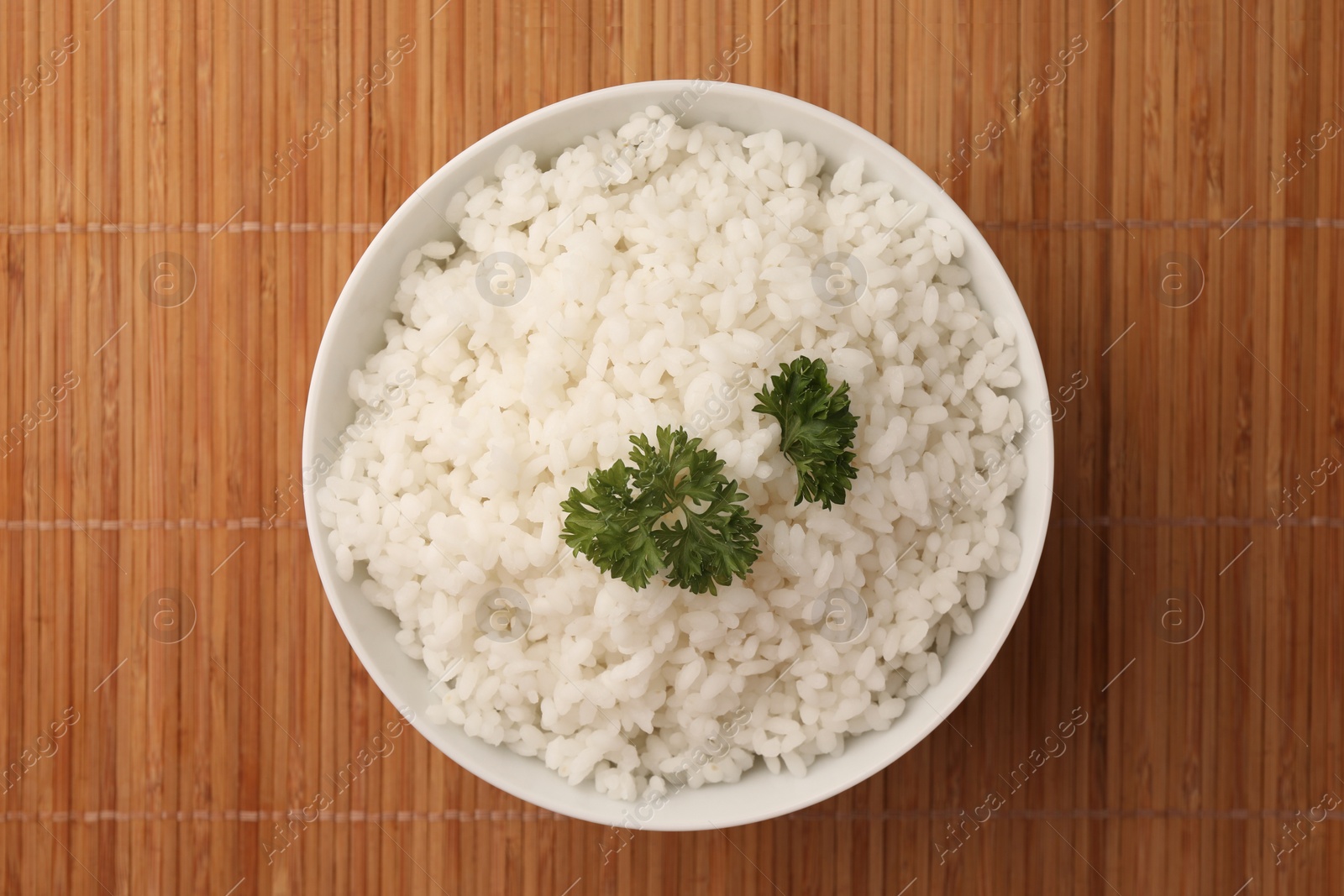 Photo of Bowl with delicious rice and parsley on bamboo mat, top view